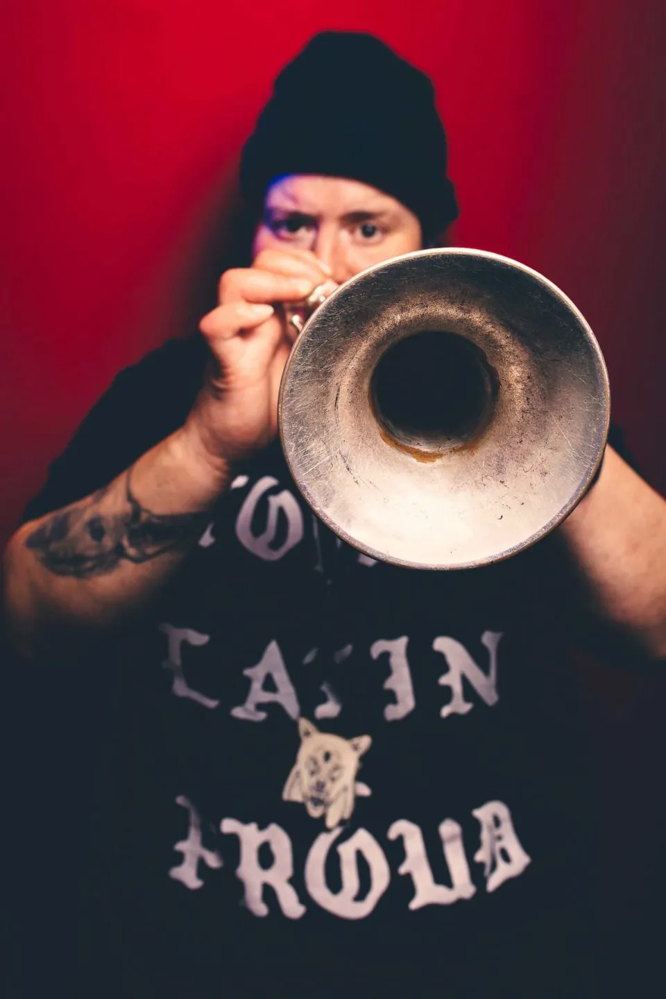 A trumpet player standing in front of a deeply-colored wall, holds their instrument up to the viewer, obscuring most of their face.